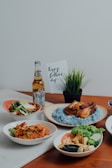 A collection of dishes is beautifully arranged on a wooden table, including a plate with blue rice and roasted chicken, a bowl with cooked prawns garnished with herbs, a bowl of mixed vegetables, and a dish with sliced omelet and fresh greens. There is a bottle of beer labeled 'Tiger Crystal,' a small potted plant, and a card that reads 'happy father's day.'
