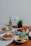 A collection of dishes is beautifully arranged on a wooden table, including a plate with blue rice and roasted chicken, a bowl with cooked prawns garnished with herbs, a bowl of mixed vegetables, and a dish with sliced omelet and fresh greens. There is a bottle of beer labeled 'Tiger Crystal,' a small potted plant, and a card that reads 'happy father's day.'