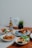 A collection of dishes is beautifully arranged on a wooden table, including a plate with blue rice and roasted chicken, a bowl with cooked prawns garnished with herbs, a bowl of mixed vegetables, and a dish with sliced omelet and fresh greens. There is a bottle of beer labeled 'Tiger Crystal,' a small potted plant, and a card that reads 'happy father's day.'