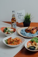 An array of dishes featuring a plate of blue rice with grilled chicken, a dish with sautéed vegetables, a bowl with prawns and garnish, and a platter with sliced tomatoes, lettuce, and a small container of green sauce. A bottle of Tiger Crystal beer is placed next to a 'Happy Father's Day' card and a small potted plant.