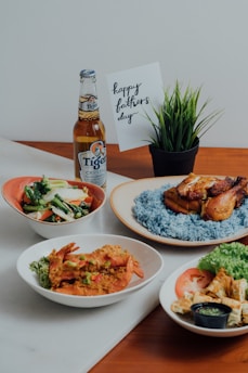 An array of dishes featuring a plate of blue rice with grilled chicken, a dish with sautéed vegetables, a bowl with prawns and garnish, and a platter with sliced tomatoes, lettuce, and a small container of green sauce. A bottle of Tiger Crystal beer is placed next to a 'Happy Father's Day' card and a small potted plant.