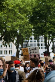A crowd of people is gathered outdoors, many wearing face masks. Several individuals hold signs, with one prominently featuring the words 'Social Justice' and a depiction of scales. Another sign references observing America. The crowd is standing close together, surrounded by greenery and buildings.