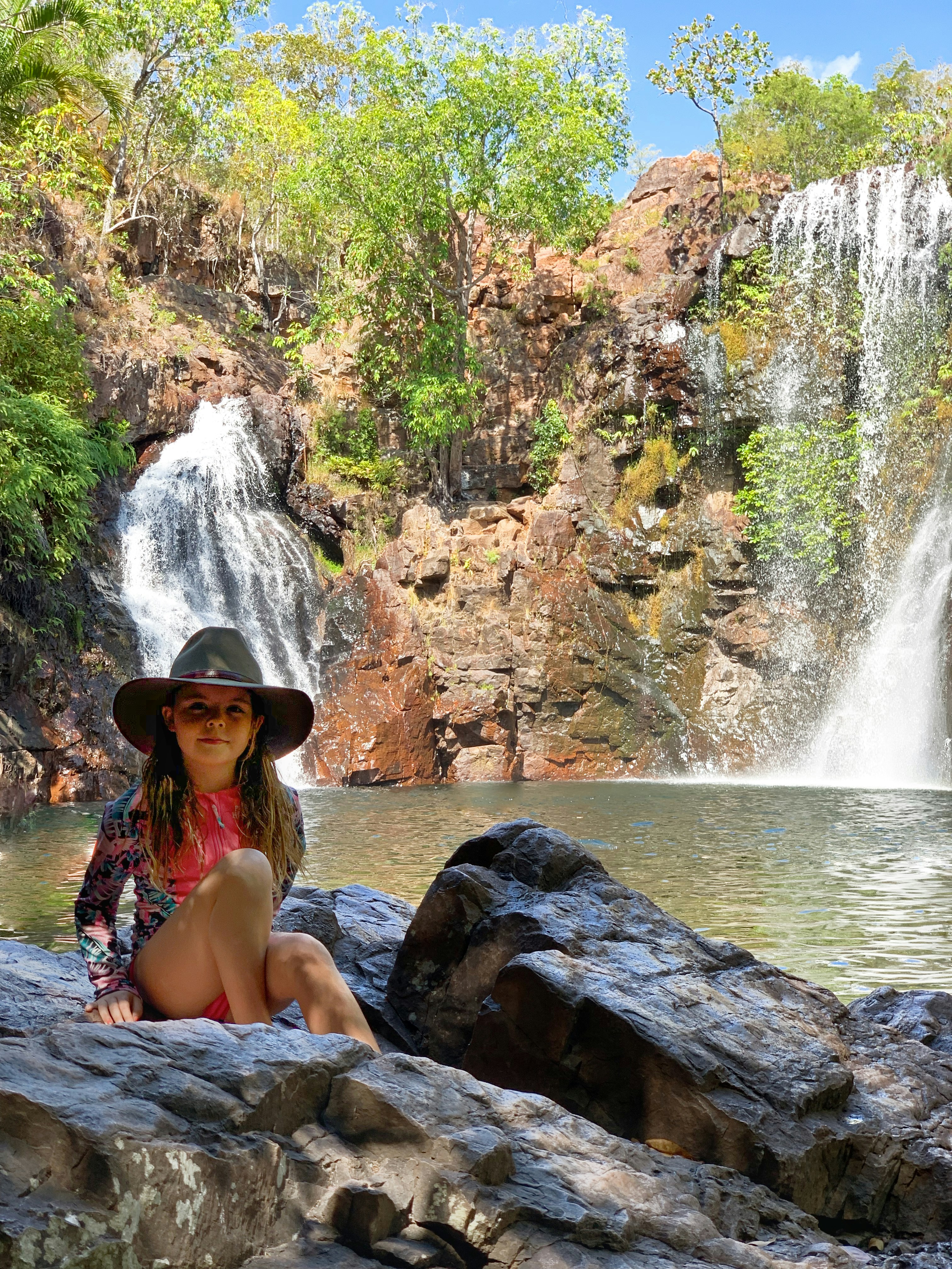 Frau im blau-weißen Bikini sitzt tagsüber auf Felsen in der Nähe von Wasserfällen