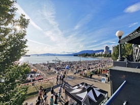 A lively beach scene with a large crowd gathered by the shoreline. People are gathered on the sand and promenade, with many sitting on blankets or chairs. In the background, the calm sea stretches out to the horizon, with mountains visible in the distance. Tall buildings line the right side of the beach, while trees border the left. The sky is bright and clear, with a few wispy clouds.