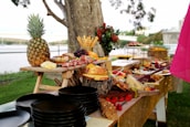 A lavish outdoor buffet spread on wooden tables features an array of gourmet items including a whole pineapple, various cheeses, cured meats, grapes, strawberries, and bread. The background displays a park-like setting with trees and a distant view of a bridge and water. Dark plates are stacked in the foreground, and decorative flowers enhance the presentation.