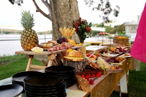 A lavish outdoor buffet spread on wooden tables features an array of gourmet items including a whole pineapple, various cheeses, cured meats, grapes, strawberries, and bread. The background displays a park-like setting with trees and a distant view of a bridge and water. Dark plates are stacked in the foreground, and decorative flowers enhance the presentation.