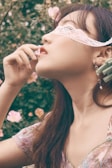 A woman with long dark hair is wearing a lace blindfold and holding a flower delicately near her lips. She appears to be dressed in a floral top. The background is filled with soft-focus greenery and blooming pink flowers, creating a serene and romantic setting.