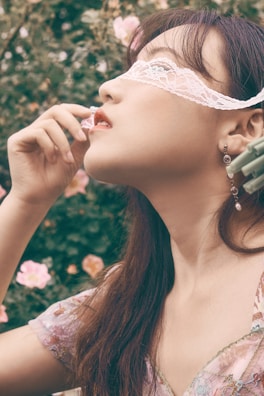 A woman with long dark hair is wearing a lace blindfold and holding a flower delicately near her lips. She appears to be dressed in a floral top. The background is filled with soft-focus greenery and blooming pink flowers, creating a serene and romantic setting.