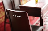 A close-up view of a wooden chair with a shiny finish beside a matching wooden table. The chair features a simple backrest with three circular cutouts. The table appears to have intricate carvings on its side, and the setting is softly lit with warm tones.