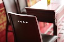 A close-up view of a wooden chair with a shiny finish beside a matching wooden table. The chair features a simple backrest with three circular cutouts. The table appears to have intricate carvings on its side, and the setting is softly lit with warm tones.
