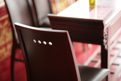 A close-up view of a wooden chair with a shiny finish beside a matching wooden table. The chair features a simple backrest with three circular cutouts. The table appears to have intricate carvings on its side, and the setting is softly lit with warm tones.