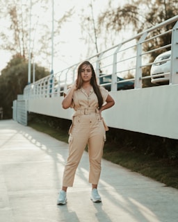 A person stands confidently on a pathway next to a railing. The individual is wearing a beige jumpsuit and white sneakers. The background includes trees and parked cars in a sunlit outdoor setting.