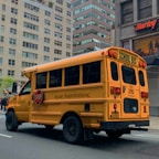 A yellow school bus parked on a city street. The bus has 'RELIANT TRANSPORTATION INC.' written on the side and features a stop sign on its side. The scene takes place in an urban environment with tall buildings in the background, including a Harley-Davidson store. The road is empty, suggesting a calm environment.