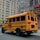 A yellow school bus parked on a city street. The bus has 'RELIANT TRANSPORTATION INC.' written on the side and features a stop sign on its side. The scene takes place in an urban environment with tall buildings in the background, including a Harley-Davidson store. The road is empty, suggesting a calm environment.