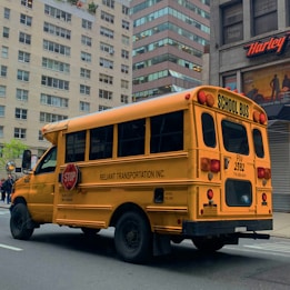 A yellow school bus parked on a city street. The bus has 'RELIANT TRANSPORTATION INC.' written on the side and features a stop sign on its side. The scene takes place in an urban environment with tall buildings in the background, including a Harley-Davidson store. The road is empty, suggesting a calm environment.