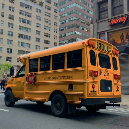 A yellow school bus parked on a city street. The bus has 'RELIANT TRANSPORTATION INC.' written on the side and features a stop sign on its side. The scene takes place in an urban environment with tall buildings in the background, including a Harley-Davidson store. The road is empty, suggesting a calm environment.