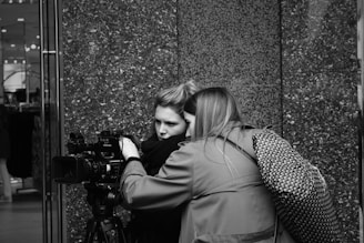 Two people are closely examining a professional video camera, possibly during a filming or photography session. They appear focused, with one person operating the camera and the other observing. The setting is urban, indicated by the smooth, textured wall in the background.