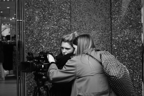 Two people are closely examining a professional video camera, possibly during a filming or photography session. They appear focused, with one person operating the camera and the other observing. The setting is urban, indicated by the smooth, textured wall in the background.