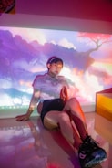 A person sits on a glossy floor, with a colorful and surreal digital projection on the wall behind them. The background resembles a dreamy landscape with soft clouds, vivid colors, and abstract forms. The individual is dressed casually, wearing shorts, a white t-shirt, and sneakers. They have glasses and short hair, appearing relaxed and thoughtful.