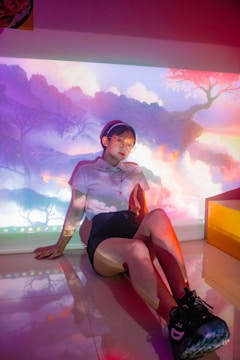 A person sits on a glossy floor, with a colorful and surreal digital projection on the wall behind them. The background resembles a dreamy landscape with soft clouds, vivid colors, and abstract forms. The individual is dressed casually, wearing shorts, a white t-shirt, and sneakers. They have glasses and short hair, appearing relaxed and thoughtful.