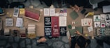 A top-down view of a marketplace stall displaying an array of books, notebooks, and art prints. The table is filled with various colorful covers and designs, including motivational phrases and illustrations. Two people are interacting at the table, possibly discussing or purchasing items.