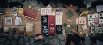 A top-down view of a marketplace stall displaying an array of books, notebooks, and art prints. The table is filled with various colorful covers and designs, including motivational phrases and illustrations. Two people are interacting at the table, possibly discussing or purchasing items.