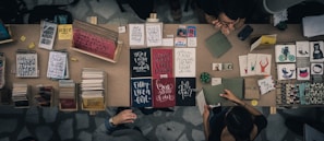 A top-down view of a marketplace stall displaying an array of books, notebooks, and art prints. The table is filled with various colorful covers and designs, including motivational phrases and illustrations. Two people are interacting at the table, possibly discussing or purchasing items.