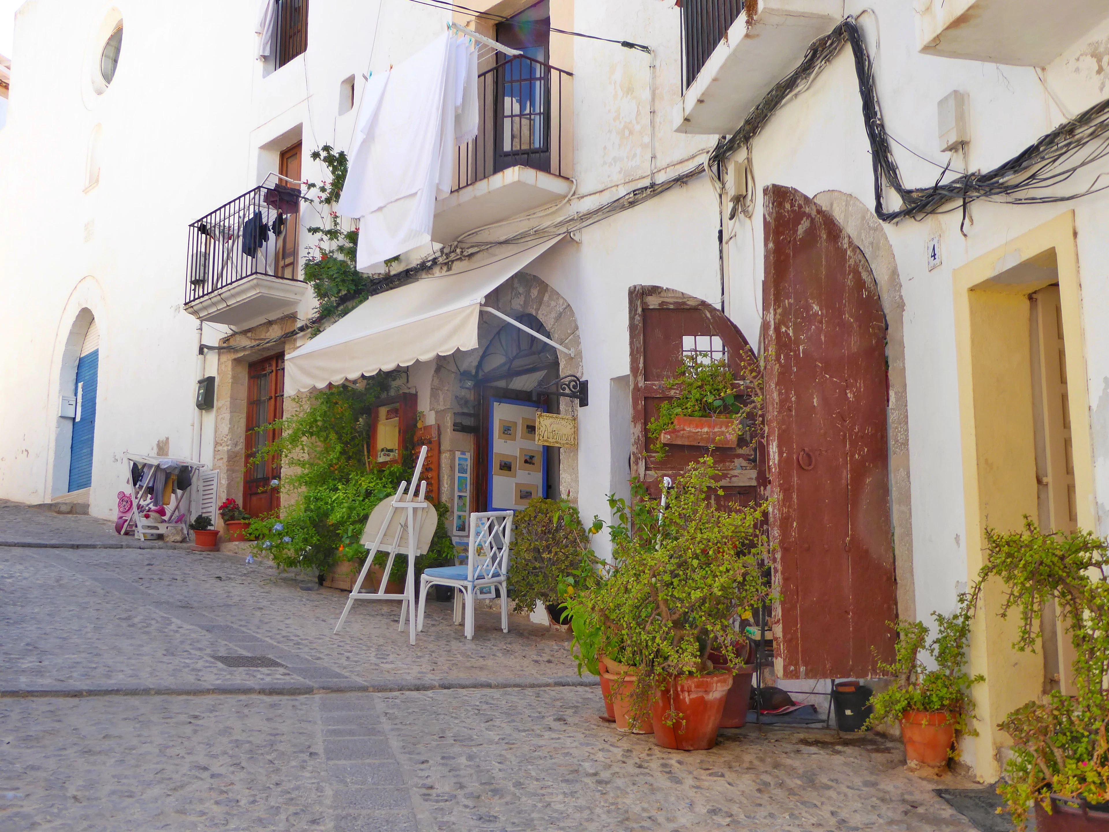 A cobbled lane in Ibiza's old town lined with potted plants, an easel and chairs outside a shaded doorway.