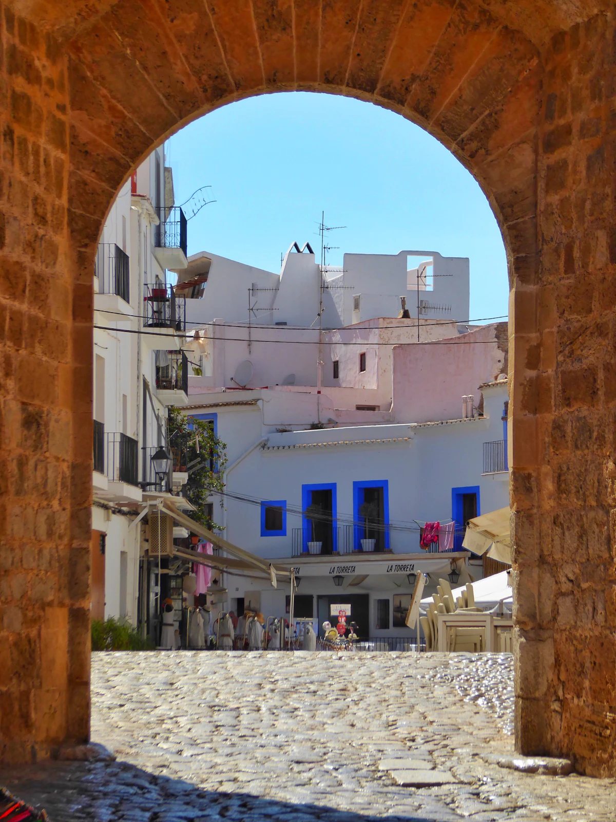 A stone arch framing whitewashed buildings with blue accents along a cobbled street in Ibiza Old Town