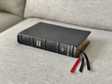 A black leather-bound book with gold lettering titled 'Holy Bible' rests on a light gray fabric sofa. Three ribbon bookmarks hanging from the book are colored red and black.