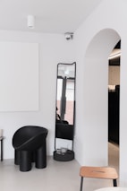 A minimalist interior with a modern black chair, a tall mirror, and a clean white wall. An archway opens to another room, and a ceiling-mounted camera is seen above. The color palette is neutral, with contrasts between black furnishings and white walls.