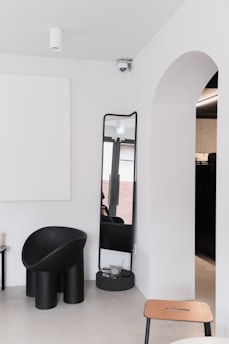 A minimalist interior with a modern black chair, a tall mirror, and a clean white wall. An archway opens to another room, and a ceiling-mounted camera is seen above. The color palette is neutral, with contrasts between black furnishings and white walls.