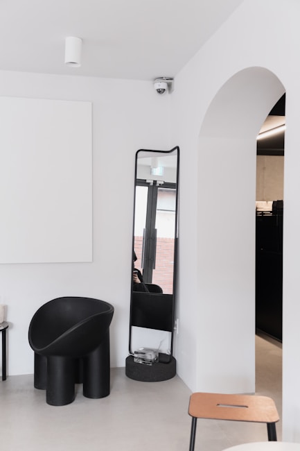 A minimalist interior with a modern black chair, a tall mirror, and a clean white wall. An archway opens to another room, and a ceiling-mounted camera is seen above. The color palette is neutral, with contrasts between black furnishings and white walls.