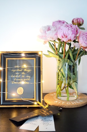 A decorative scene featuring a framed quote with elegant gold lettering surrounded by fairy lights, alongside a vase of pink peonies on a cork coaster. Some bookmarks or cards are laid on the table.