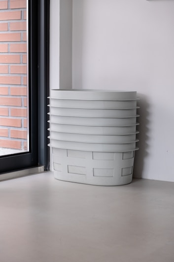 A stack of white plastic baskets is neatly positioned in the corner of a room. The background includes a large window on the left with brickwork visible outside, and the walls are plain and white.