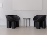 Two modern, black armchairs with curved designs stand on either side of a small, round, black side table. The setting has a minimalist aesthetic with white walls and a light gray floor, creating a stark contrast with the dark furniture.