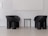 Two modern, black armchairs with curved designs stand on either side of a small, round, black side table. The setting has a minimalist aesthetic with white walls and a light gray floor, creating a stark contrast with the dark furniture.