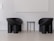 Two modern, black armchairs with curved designs stand on either side of a small, round, black side table. The setting has a minimalist aesthetic with white walls and a light gray floor, creating a stark contrast with the dark furniture.