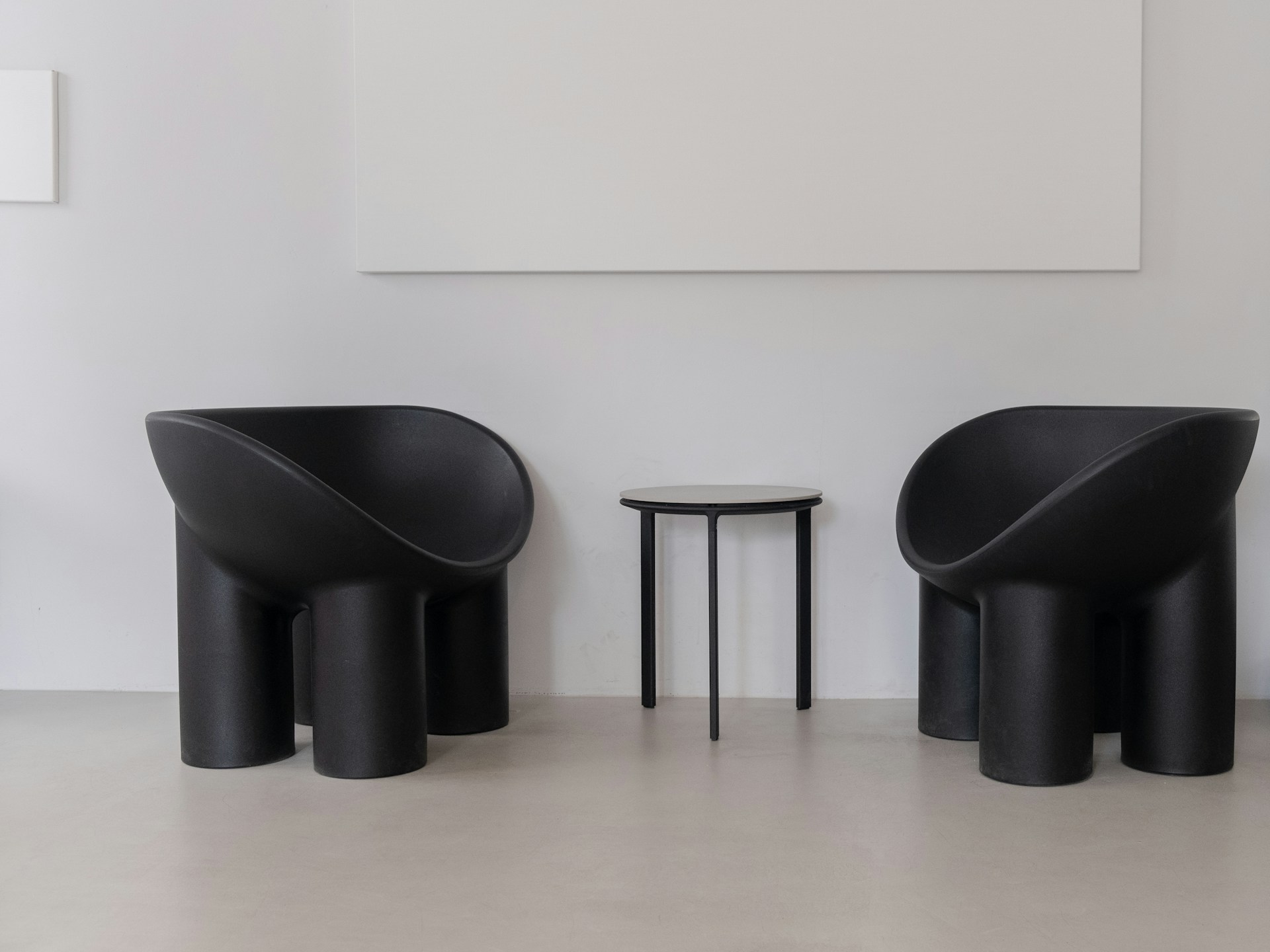 Two modern, black armchairs with curved designs stand on either side of a small, round, black side table. The setting has a minimalist aesthetic with white walls and a light gray floor, creating a stark contrast with the dark furniture.