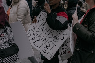 Several people are holding signs and gathering in a protest. One sign prominently reads 'Witches Stand Against Racism' with other signs partially visible. Participants are dressed in warm clothing.