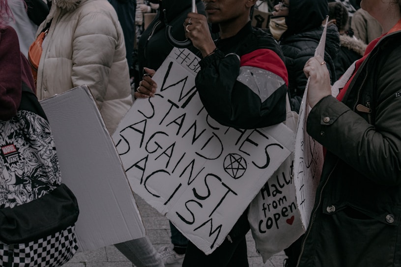 Several people are holding signs and gathering in a protest. One sign prominently reads 'Witches Stand Against Racism' with other signs partially visible. Participants are dressed in warm clothing.
