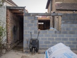 A partially constructed brick wall stands with an opening resembling a window. In front of the wall, a black wheelbarrow is placed alongside a shovel. The surrounding area appears to be a construction site with various building materials, such as a bag of cement, scattered around. Visible are older brick structures to the left and right, suggesting an ongoing building extension.