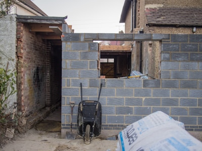 A partially constructed brick wall stands with an opening resembling a window. In front of the wall, a black wheelbarrow is placed alongside a shovel. The surrounding area appears to be a construction site with various building materials, such as a bag of cement, scattered around. Visible are older brick structures to the left and right, suggesting an ongoing building extension.