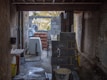 A construction site with unfinished walls made of concrete blocks and bricks. Various construction materials and tools are scattered around, including a cement mixer and a stack of bricks. The area is dimly lit, with natural light filtering in from openings in the unfinished structure.
