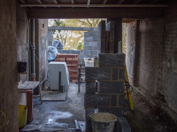 A construction site with unfinished walls made of concrete blocks and bricks. Various construction materials and tools are scattered around, including a cement mixer and a stack of bricks. The area is dimly lit, with natural light filtering in from openings in the unfinished structure.
