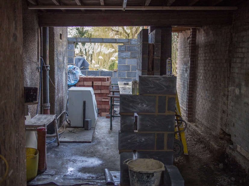 A construction site with unfinished walls made of concrete blocks and bricks. Various construction materials and tools are scattered around, including a cement mixer and a stack of bricks. The area is dimly lit, with natural light filtering in from openings in the unfinished structure.