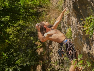 A man is engaged in rock climbing on a rugged cliff face, surrounded by lush greenery. His upper body is exposed, showing the physical exertion and focus required for the climb. He is wearing climbing gear, including a harness, and is reaching for a higher point on the cliff.