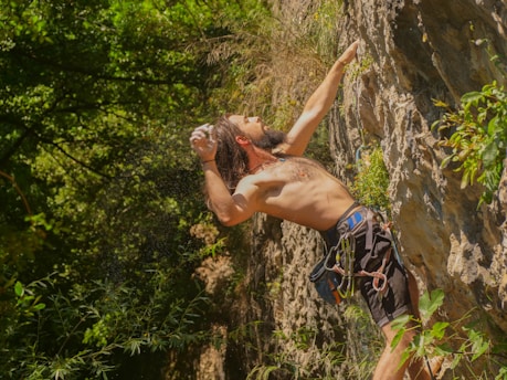A man is engaged in rock climbing on a rugged cliff face, surrounded by lush greenery. His upper body is exposed, showing the physical exertion and focus required for the climb. He is wearing climbing gear, including a harness, and is reaching for a higher point on the cliff.