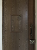A wooden door with a prominent wood grain texture features a small square hatch in the center. The latch of the hatch is visible, designed for securing it shut. On the right side of the door, a vintage-style metal lock is installed along with an intricate handle.