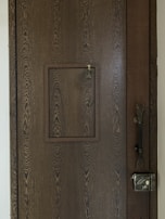 A wooden door with a prominent wood grain texture features a small square hatch in the center. The latch of the hatch is visible, designed for securing it shut. On the right side of the door, a vintage-style metal lock is installed along with an intricate handle.